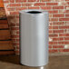 A silver Rubbermaid Atrium waste receptacle next to a brick wall.