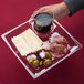 A person holding a WNA Comet clear plastic square cocktail plate with a glass of wine and food.
