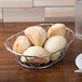An American Metalcraft oval chrome wire basket filled with bread rolls on a table.