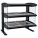 A black and silver Hatco countertop heated zone merchandiser with three shelves holding trays on a counter.