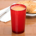 A red Cambro plastic tumbler filled with orange liquid on a table with a plate of bagels.