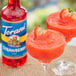 A glass of red drink with a strawberry on the rim next to a bottle of Torani Sugar-Free Strawberry Fruit Syrup.