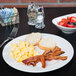 A Tuxton AlumaTux Pearl White plate with bacon, eggs, and toast with a bowl of fruit on a table.