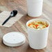 Two white Choice paper food cups with vented paper lids on a table with a cup of soup and a spoon.