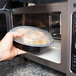A hand holding a Pactiv Newspring black plastic container of food in a microwave.