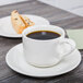 A cup of coffee on a Tuxton San Marino AlumaTux Pearl White saucer with food on a table.