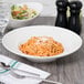 A bowl of spaghetti with sauce and cheese, a salad, and a glass of wine on a table set with a Tuxton San Marino AlumaTux Pearl White bowl.