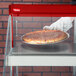 A gloved hand places a pizza on a tray in a red Hatco countertop display case.