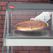 A gloved hand holds a pizza in a Hatco countertop glass case.