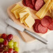 A Franmara stainless steel soft cheese knife on a cutting board with cheese and meat.