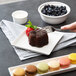 A hand holding a fork to a chocolate cake on a white 10 Strawberry Street square porcelain plate with fruit.