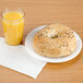 A bagel on a plate next to a Cambro clear plastic tumbler filled with orange juice.