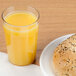A Cambro clear plastic tumbler filled with orange juice next to a plate with a bagel on it.