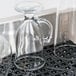 A clear glass jug with a handle on a San Jamar black interlocking bar mat with glass cups.