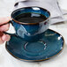 A hand holding a Tuxton Artisan Night Sky saucer with a cup of coffee on it.