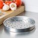 An American Metalcraft heavy weight aluminum perforated pizza pan with a white background next to garlic and tomatoes.