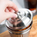 A hand holding an American Metalcraft mini stainless steel trash can lid over a metal canister.