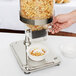 A hand pours cereal from a Vollrath cereal dispenser into a bowl on a table.