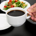 A person's hand holding a white Arcoroc Candour porcelain cup of coffee over a plate of food.