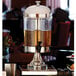 An American Metalcraft stainless steel beverage dispenser on a table with a glass container of liquid in it.