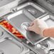 A hand in a glove picking up a Choice stainless steel steam table pan cover with food inside.