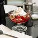 An Arcoroc glass dessert bowl with red liquid, whipped cream, and nuts on a white table with a fork.