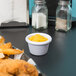 A white Carlisle ramekin filled with yellow mustard on a table with fried chicken and fries.