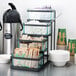 A black Cal-Mil stair step condiment display with glass jars on a counter with a coffee maker and coffee containers.