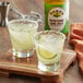 Two glasses of Rose's sweetened lime juice on a wooden board with a drink and a lime slice.