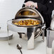 A person in gloves holding a Vollrath New York Retractable Dripless Round Chafer with brass trim full of food.