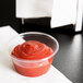 A Fabri-Kal Greenware clear plastic souffle cup with ketchup in it on a table.
