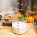 A Cambro clear polycarbonate measuring cup on a counter with a bowl of fruit filled with milk.