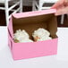 A hand holding a pink bakery box with a cupcake inside.