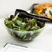 A clear Fineline PET plastic bowl filled with salad on a table with black plastic utensils.