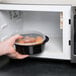 A hand holding a Pactiv Newspring black plastic VERSAtainer filled with food in a microwave.