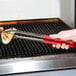 A person using Vollrath stainless steel tongs with red Kool Touch handles to grill food.