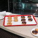 A red Carlisle Glasteel bakery tray of chocolate covered doughnuts on a counter.