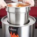 A person using a Vollrath Cayenne adapter ring to hold a pot of soup over flames on a counter.