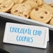 A white rectangular ceramic card sign with blue writing on it displayed on a plate of chocolate chip cookies.