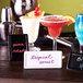 A rectangular white ceramic card sign with pink writing on it sitting on a table with a cocktail in a glass.