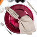 A Tabletop Classics by Walco raspberry charger plate with silverware and a napkin on it.