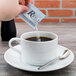 A hand pouring a Regal Blue aspartame sugar substitute packet into a white cup of coffee.