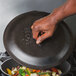 A hand holding a Lodge cast iron lid over a pan of vegetables.