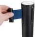 A hand holding a blue retractable belt attached to a black Aarco crowd control stanchion.