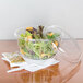 A Dart clear plastic container of salad on a table with a fork and knife.