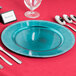 A table set with a blue Charge It by Jay plastic charger plate, silverware, and a glass.