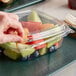 A hand picking fruit from a Dart ClearPac plastic container on a table with salad bar items.