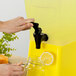 A hand using a Choice black faucet to pour lemon juice into a yellow dispenser.