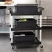 A black Cambro utility cart with three shelves holding black containers.