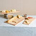 An American Metalcraft olive wood serving board with cheese, bread, and crackers on a table with food.